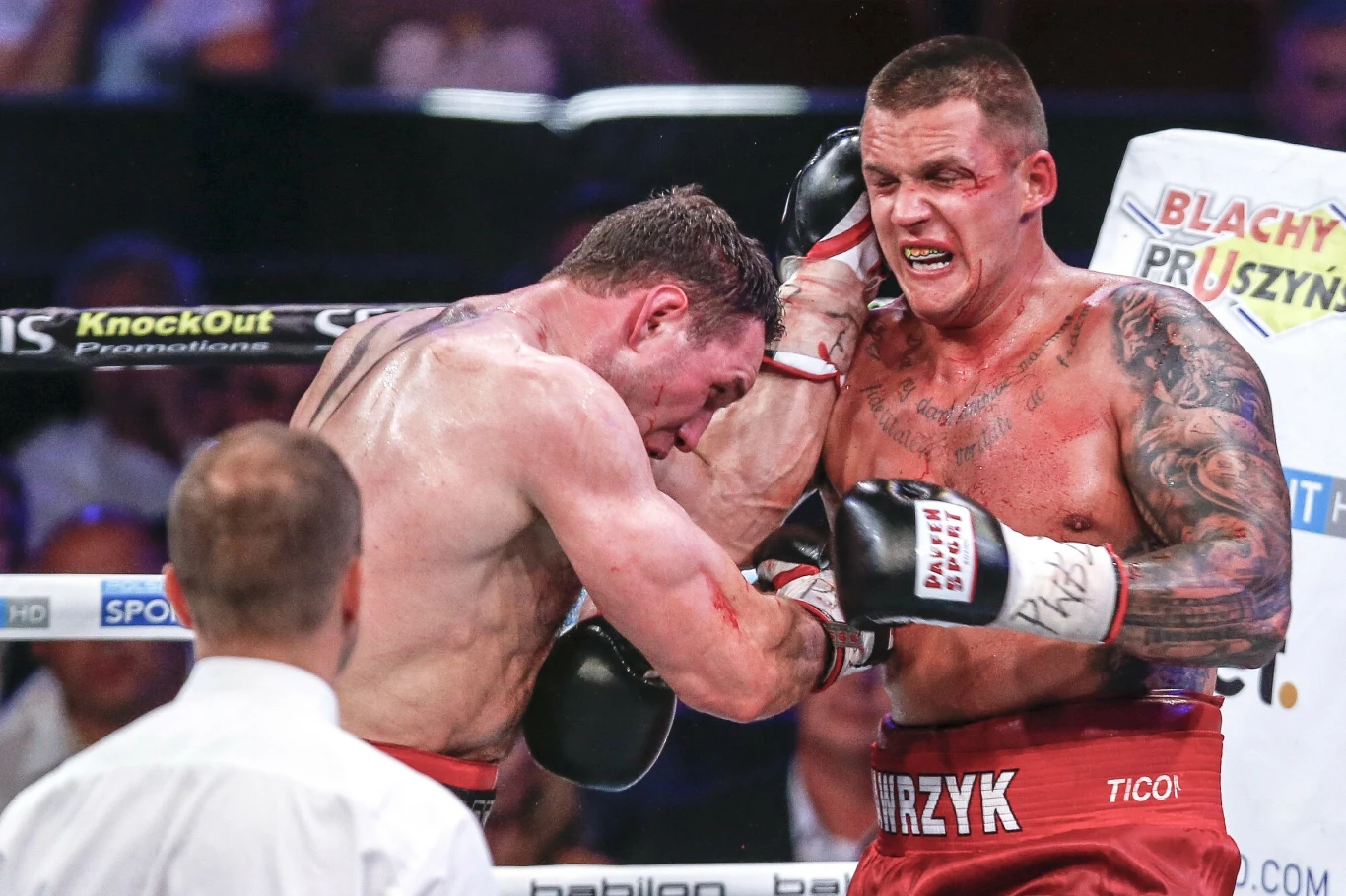 Dwóch bokserów walczy w ringu, jeden z nich mocno atakuje drugiego, który jest oparty o liny, wokół nich widoczne są ślady krwi oraz sędzia obserwujący przebieg pojedynku.