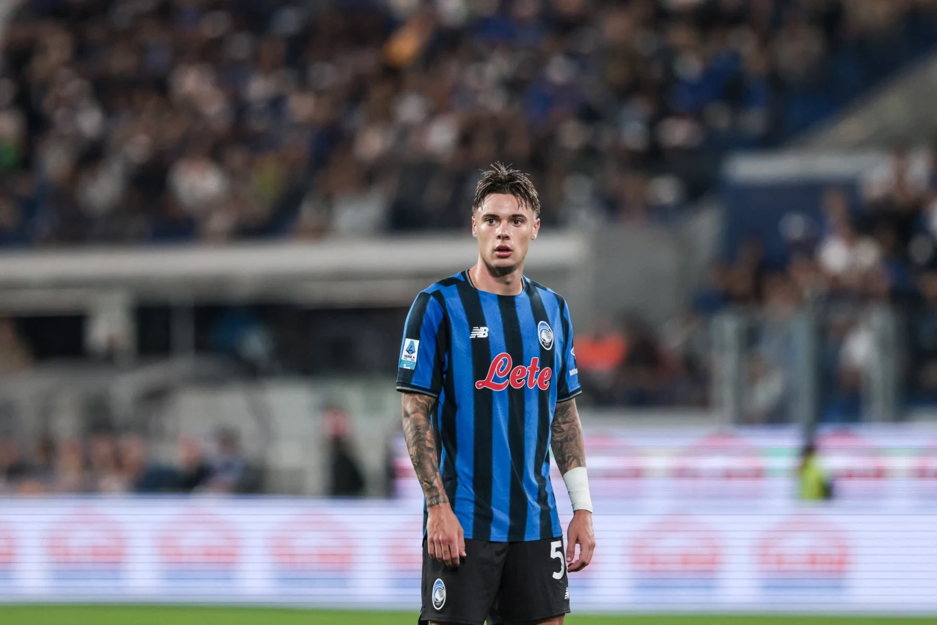 Piłkarz w czarno-niebieskiej koszulce z widocznymi tatuażami na przedramionach stoi na stadionie, wokół niego rozmazane sylwetki kibiców i trybuny.