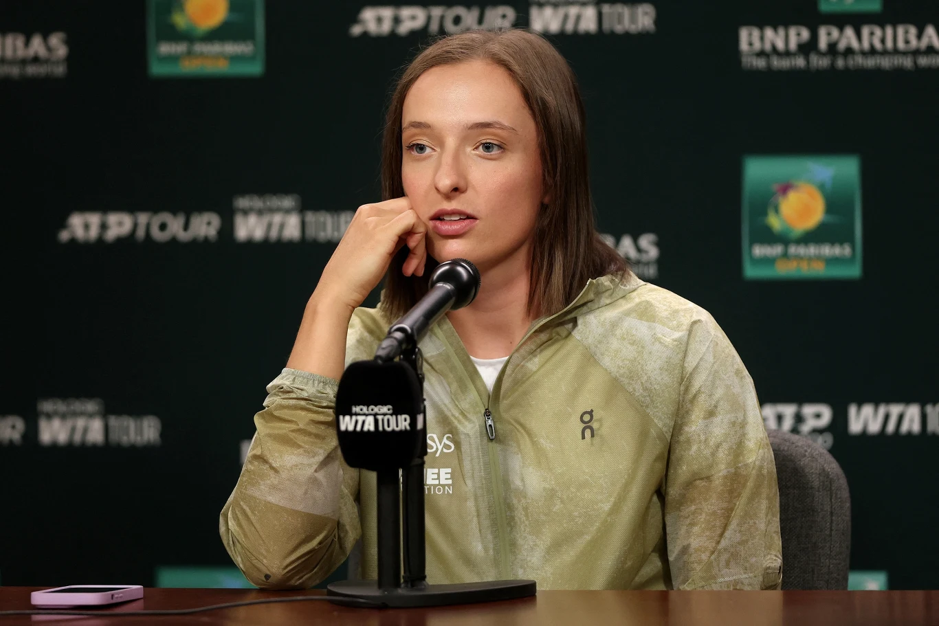 Kobieta siedząca przy stole podczas konferencji prasowej, trzymająca mikrofon, w tle logo WTA Tour oraz BNP Paribas, sportowe ubranie