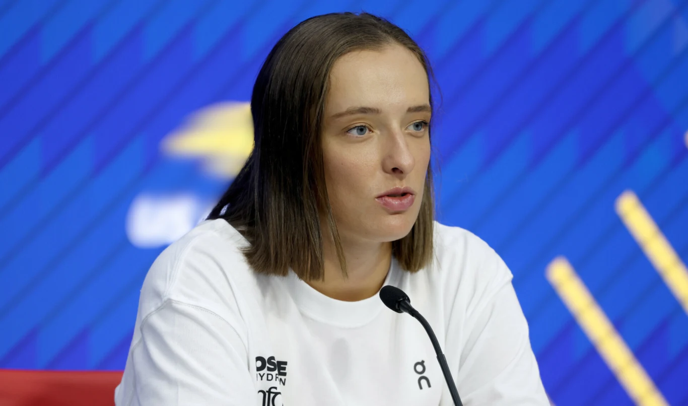 Młoda kobieta o prostych, brązowych włosach udziela wywiadu lub bierze udział w konferencji prasowej, siedząc przy mikrofonie na tle niebieskiej ściany ze wzorami i logotypami.