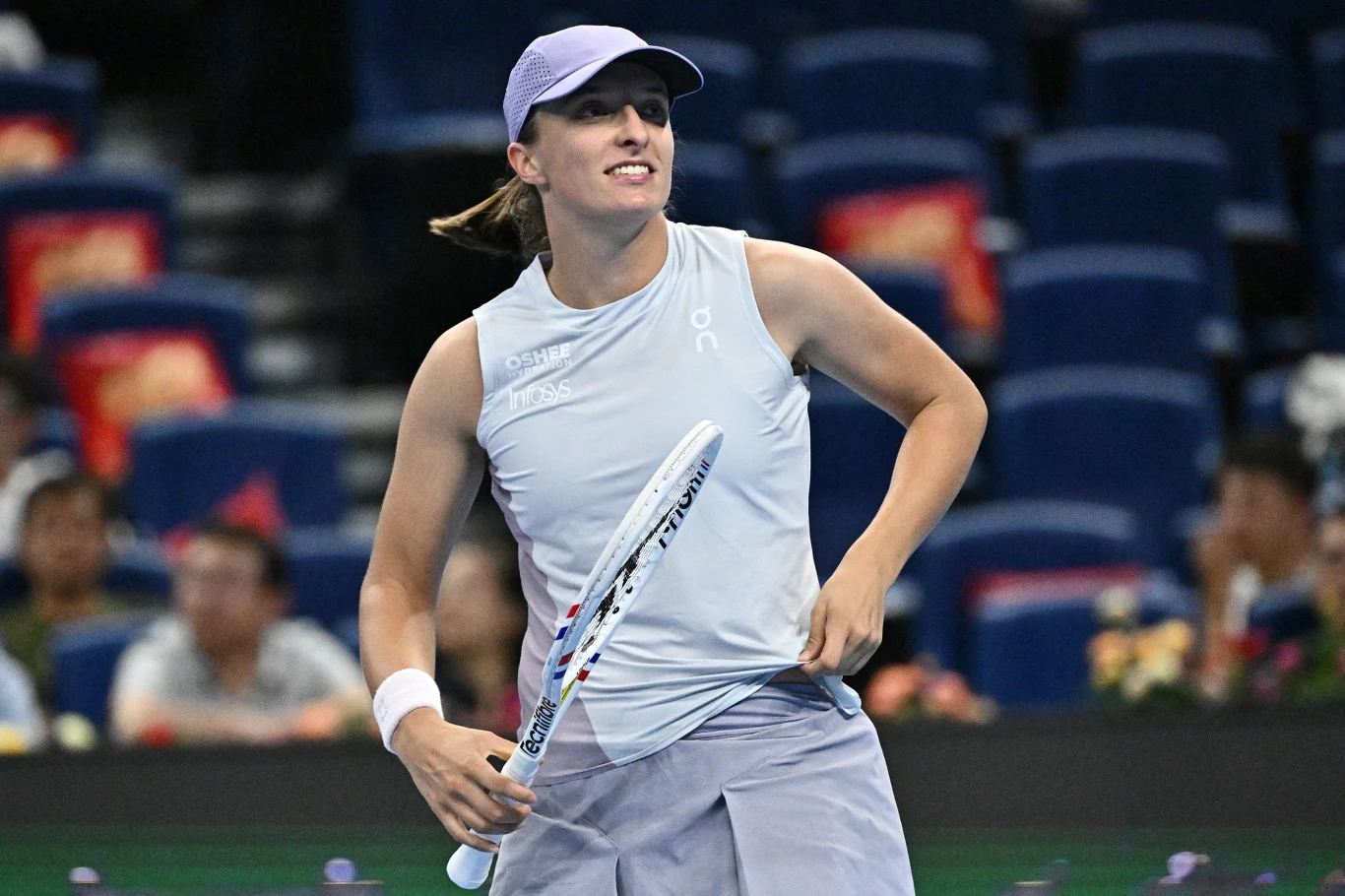 Tenisistka w sportowym stroju trzymająca rakietę na korcie podczas meczu, w tle rozmazana publiczność i niebieskie trybuny.