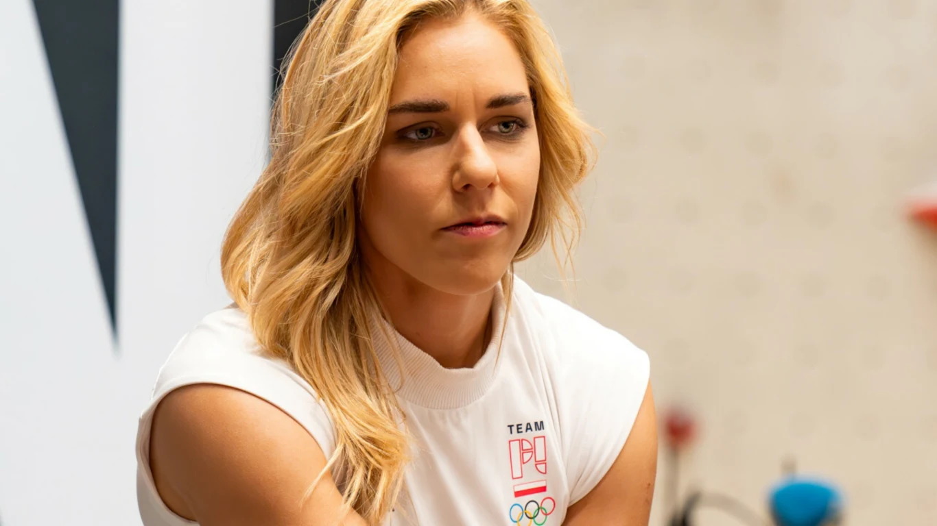 Aleksandra Mirosław Kobieta o blond włosach w białej koszulce sportowej z logo polskiej drużyny olimpijskiej, siedząca w nowoczesnym wnętrzu, skoncentrowana i zamyślona.