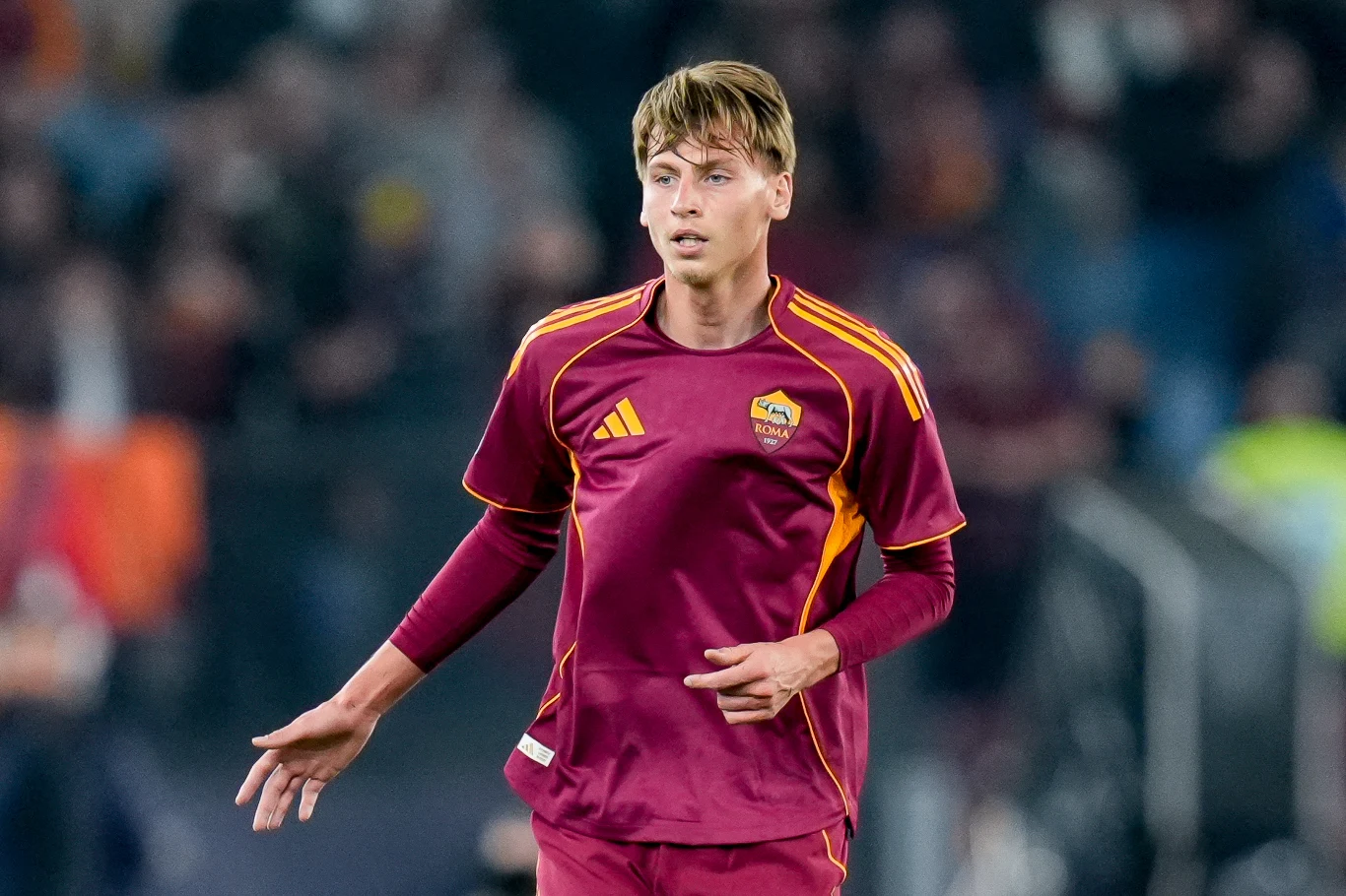 Młody piłkarz ubrany w bordowo-żółty strój drużyny AS Roma podczas meczu na stadionie, na tle rozmytej publiczności.