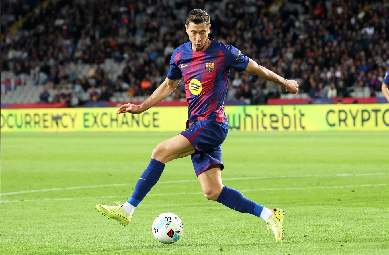 Robert Lewandowski w barwach FC Barcelona w sezonie 2025/2026 Piłkarz w stroju FC Barcelony wykonuje dynamiczne kopnięcie piłki na boisku podczas meczu, w tle widoczna trybuna pełna kibiców oraz reklamy na bandach.