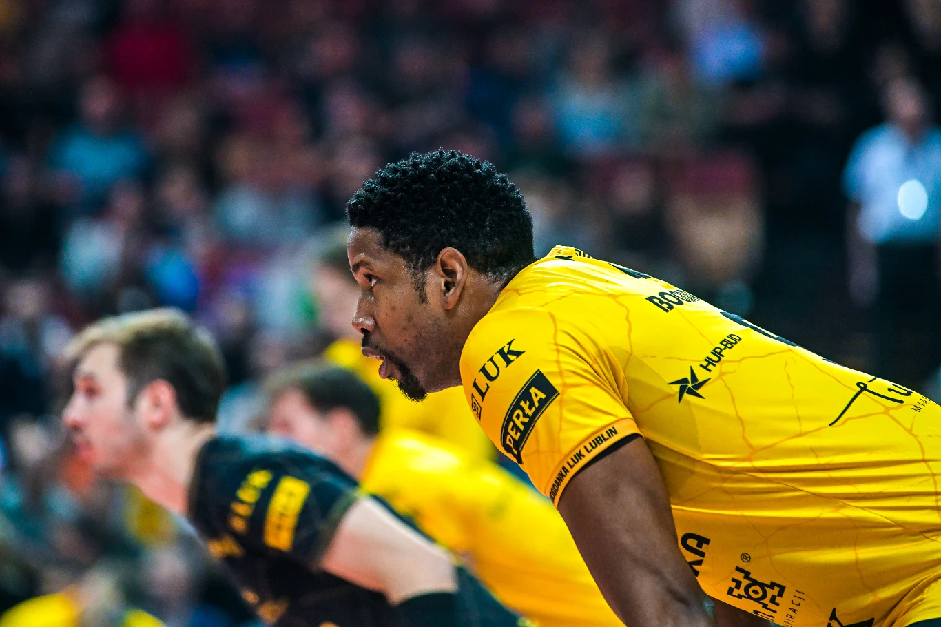 Wilfredo Leon Zawodnik drużyny piłki ręcznej w żółtej koszulce z logo sponsorów obserwuje wydarzenia na boisku, skupiony na grze, z rozmytym tłem i innymi graczami w tle.