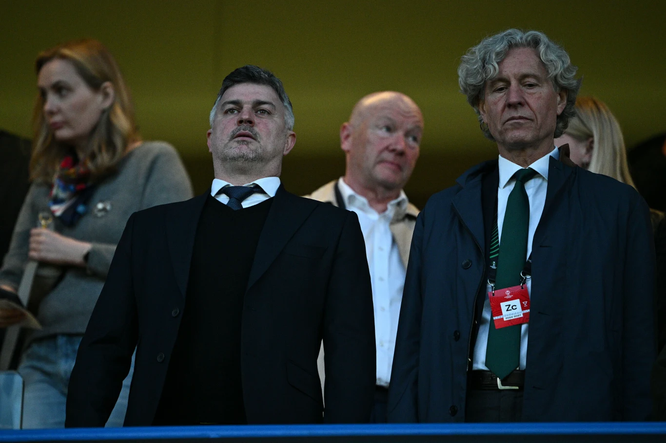 Grupa czterech osób na trybunach, dwaj mężczyźni na pierwszym planie ubrani elegancko, jeden z nich z identyfikatorem na szyi, w tle dwie osoby obserwujące wydarzenie.