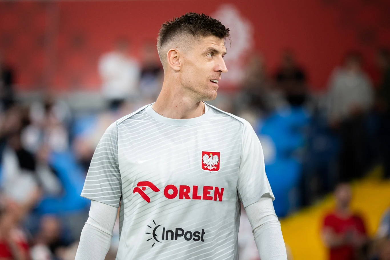 Piłkarz w białej koszulce treningowej z logo reprezentacji Polski oraz sponsorami, stojący na tle rozmytej publiczności i stadionu.