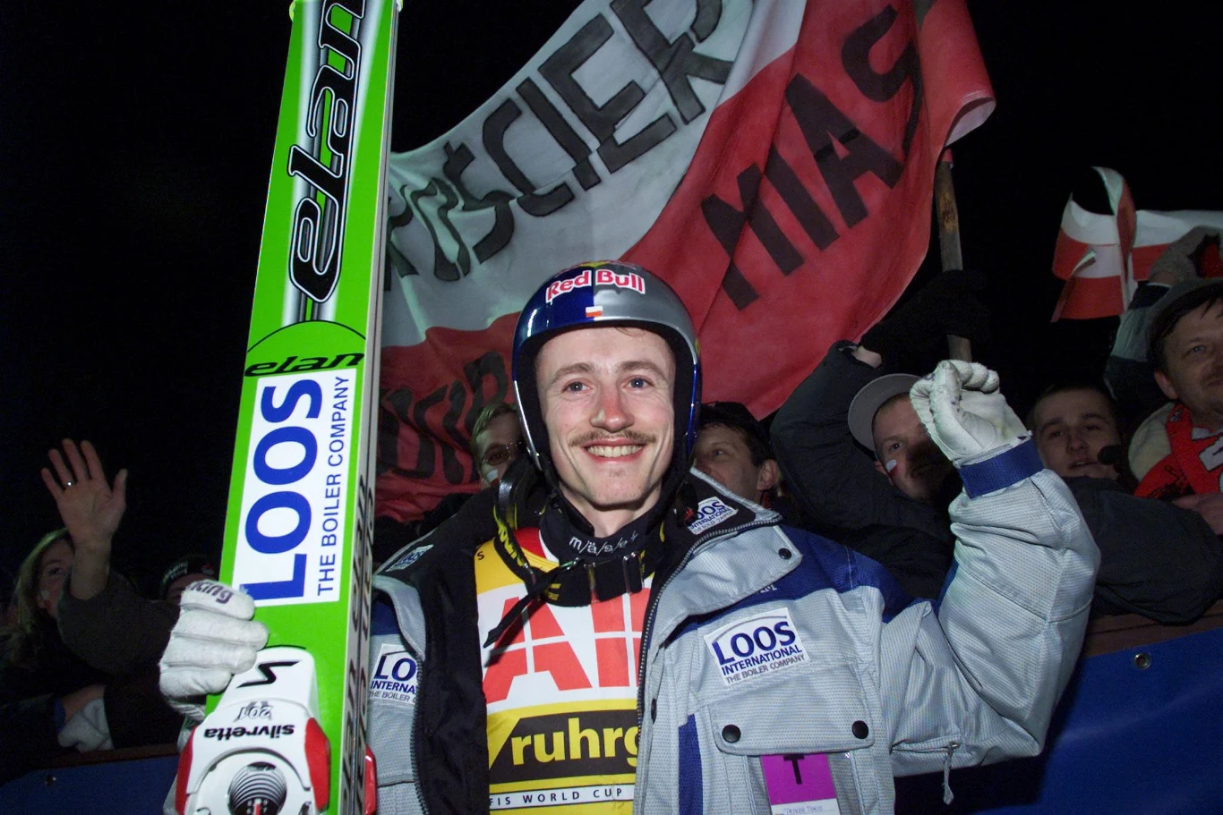 Sportowiec w stroju narciarskim trzyma narty i kask, uśmiechając się oraz unosząc zaciśniętą pięść w geście triumfu. W tle tłum kibiców z polskim transparentem oraz flagami, wyrażający radość i wsparcie.