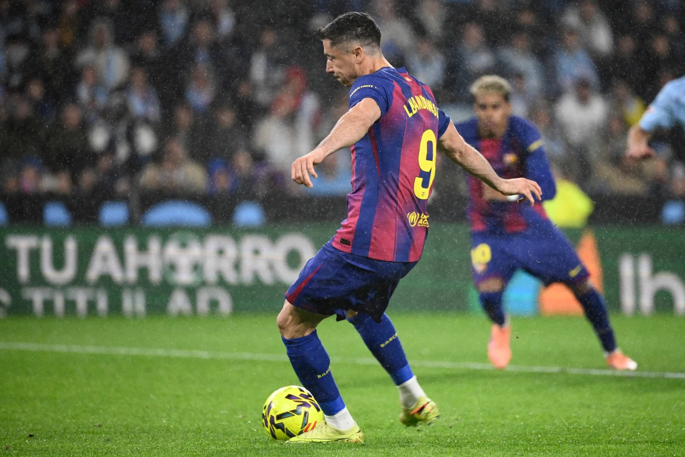 Piłkarz w koszulce Barcelony z numerem 9 kopie piłkę podczas meczu, w tle widoczny inny zawodnik oraz rozmyte trybuny.