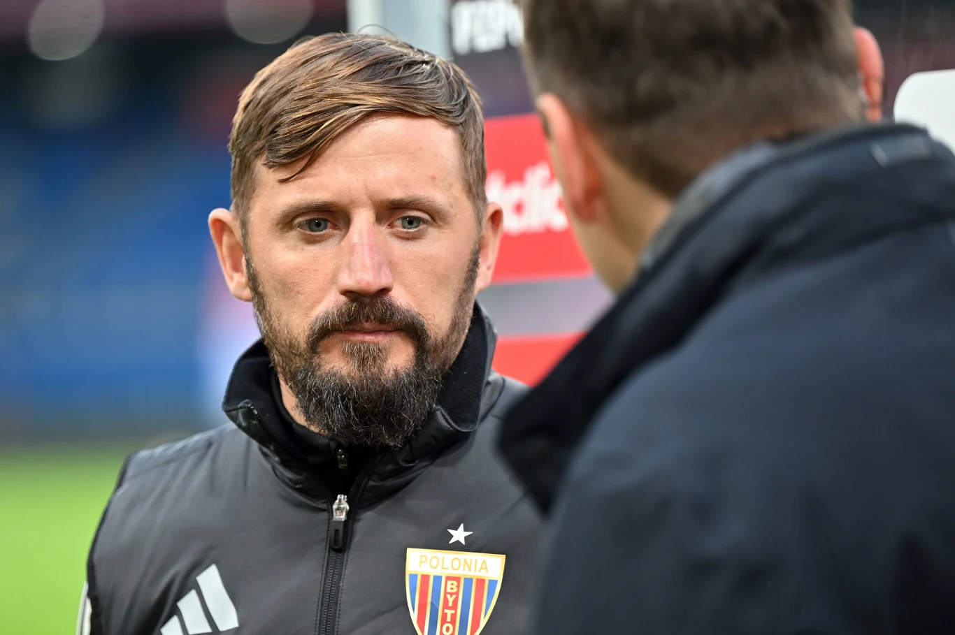 Mężczyzna z krótką fryzurą i brodą w sportowej kurtce z emblematem klubu Polonia Bytom, stojący naprzeciw drugiej osoby, która widoczna jest od tyłu i rozmawiający z nią na tle rozmytego stadionu.