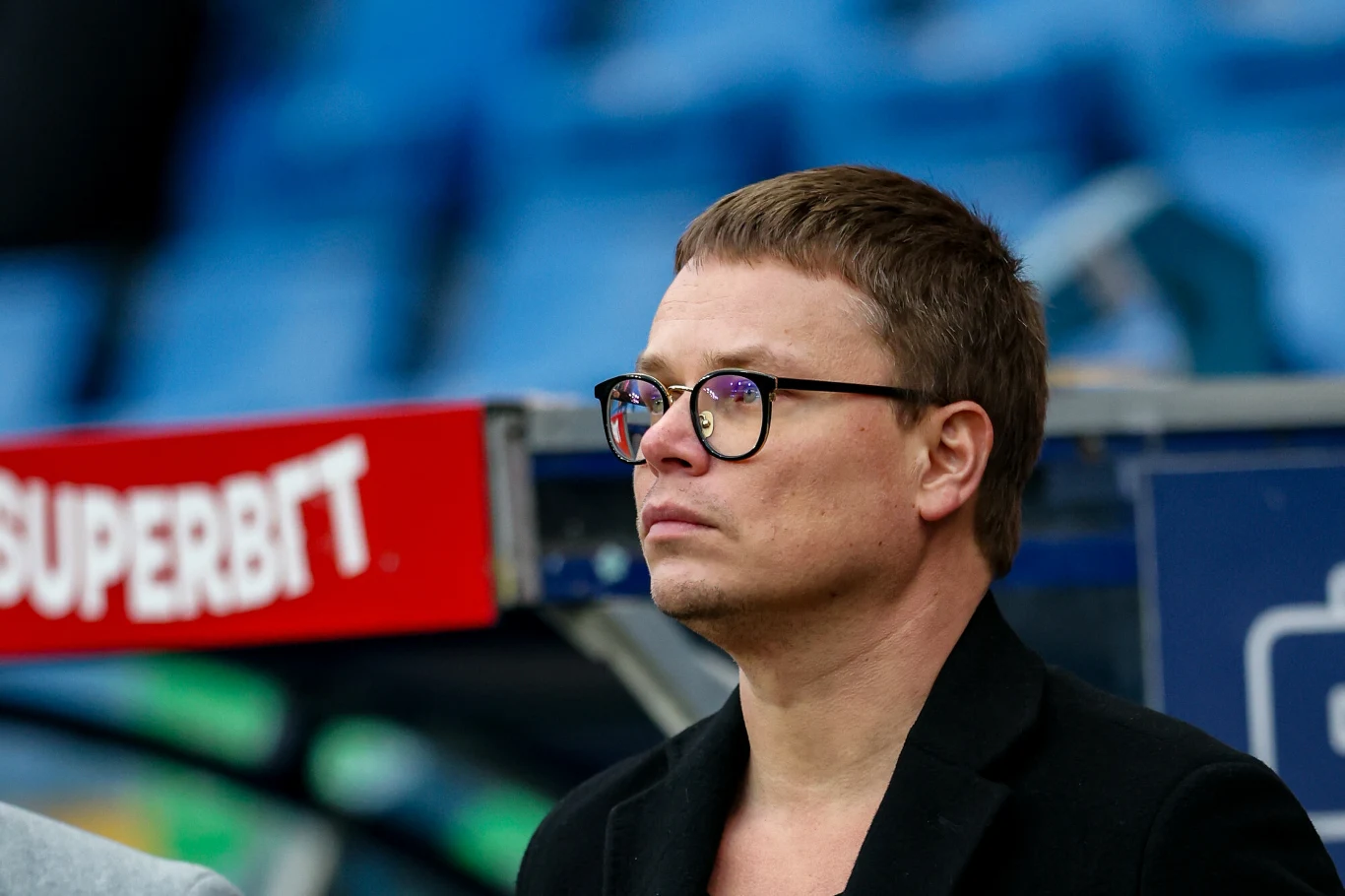 Mężczyzna w okularach i czarnej marynarce patrzy przed siebie podczas wydarzenia sportowego, w tle rozmazane niebieskie trybuny stadionu i fragment czerwonego baneru reklamowego.