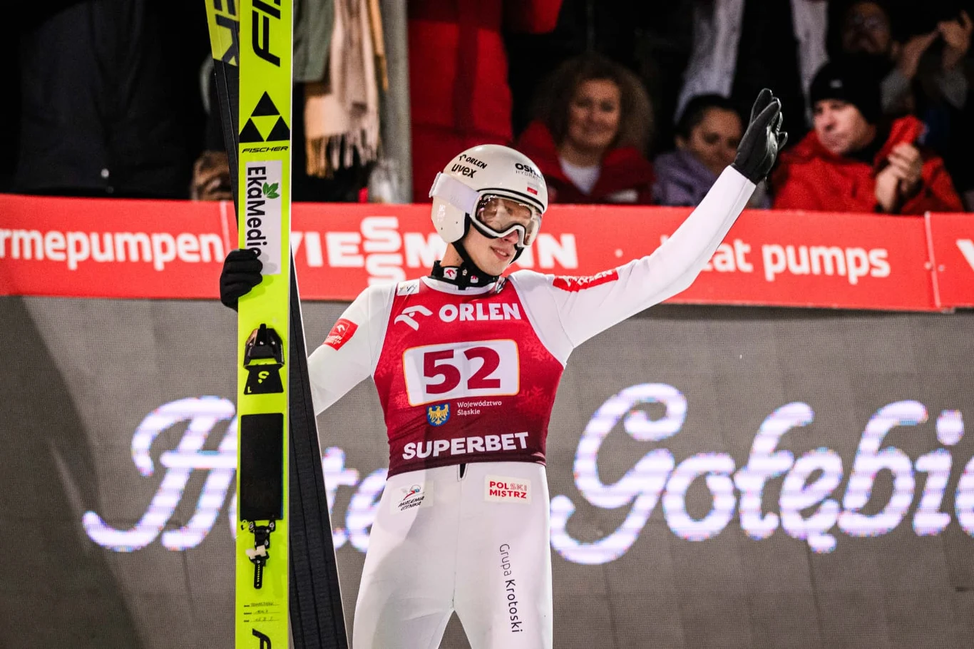 Zawodnik skoków narciarskich w pełnym stroju sportowym stoi z uniesioną ręką, trzymając narty. W tle widoczna publiczność oraz bannery reklamowe.