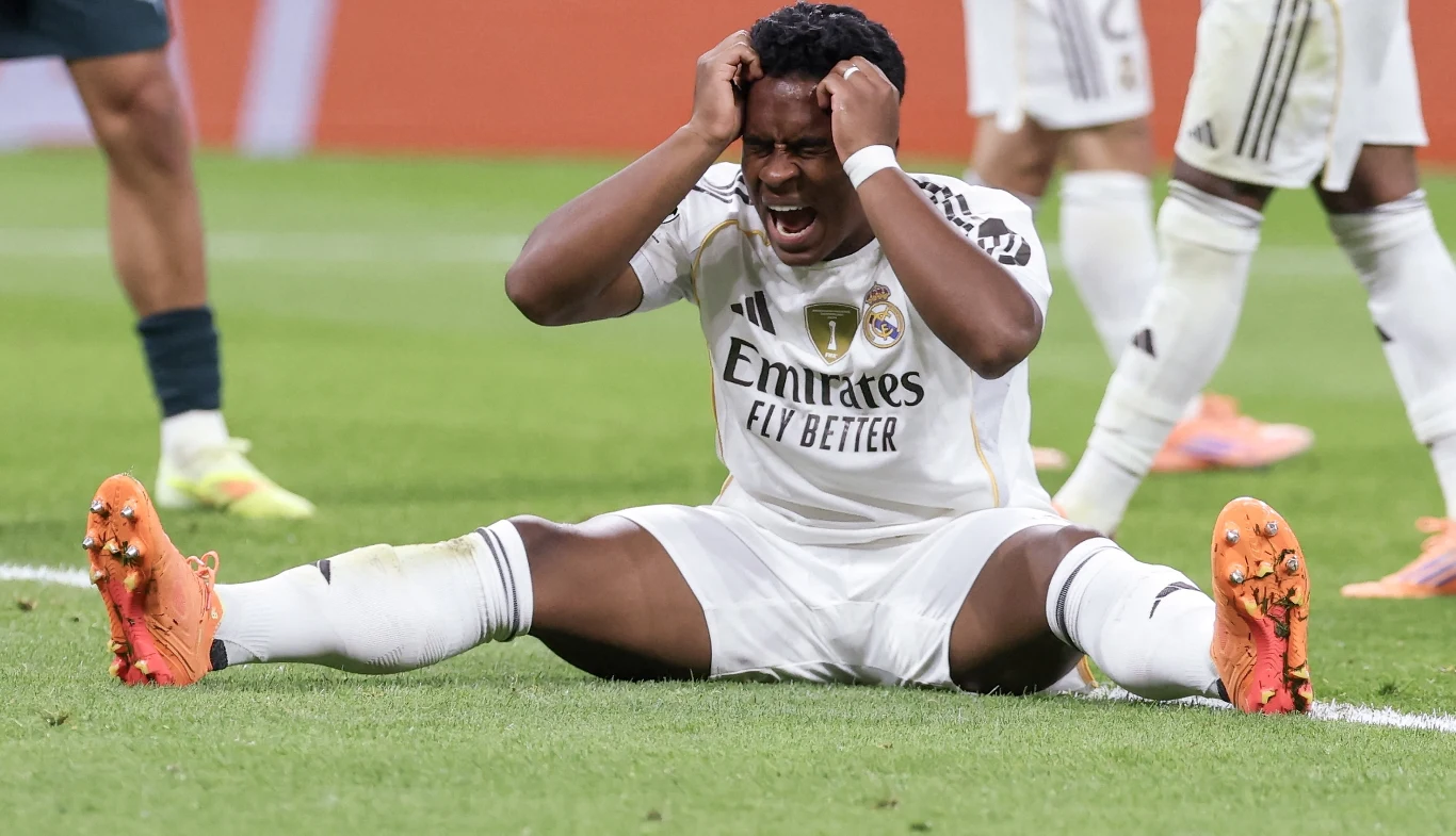 Piłkarz w stroju Realu Madryt siedzi na murawie boiska z szeroko rozstawionymi nogami, trzymając się za głowę i wyrażając frustrację lub rozczarowanie, w tle widoczne nogi innych zawodników.