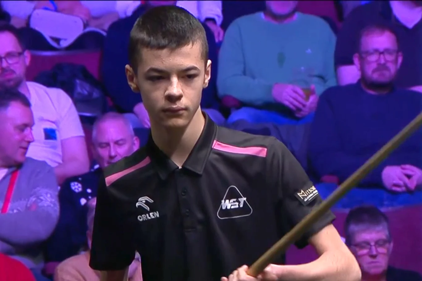 Młody zawodnik snookera w czarnej koszulce z logo sponsora trzyma kij bilardowy, za nim widownia skupiona na rozgrywce.