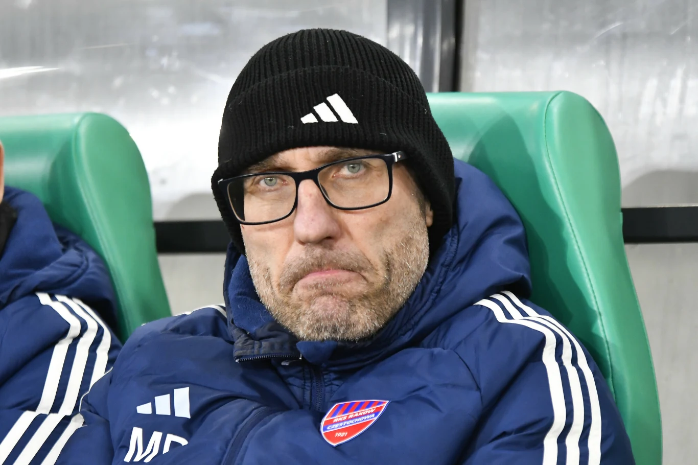 Marek Papszun Mężczyzna w okularach i czarnej czapce siedzi na zielonym fotelu stadionowym, ubrany w niebieską sportową kurtkę z herbem klubu sportowego, sprawia poważne wrażenie.