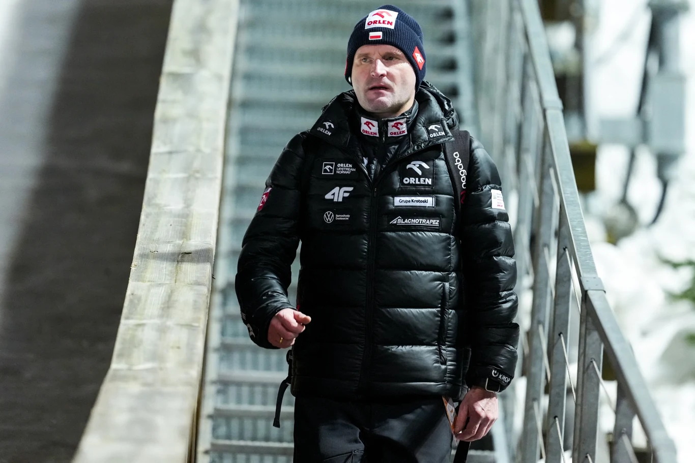 Mężczyzna w sportowej kurtce z logotypami sponsorów idzie po schodach, na głowie ma czapkę zimową, w tle widoczna metalowa barierka oraz fragment skoczni narciarskiej.