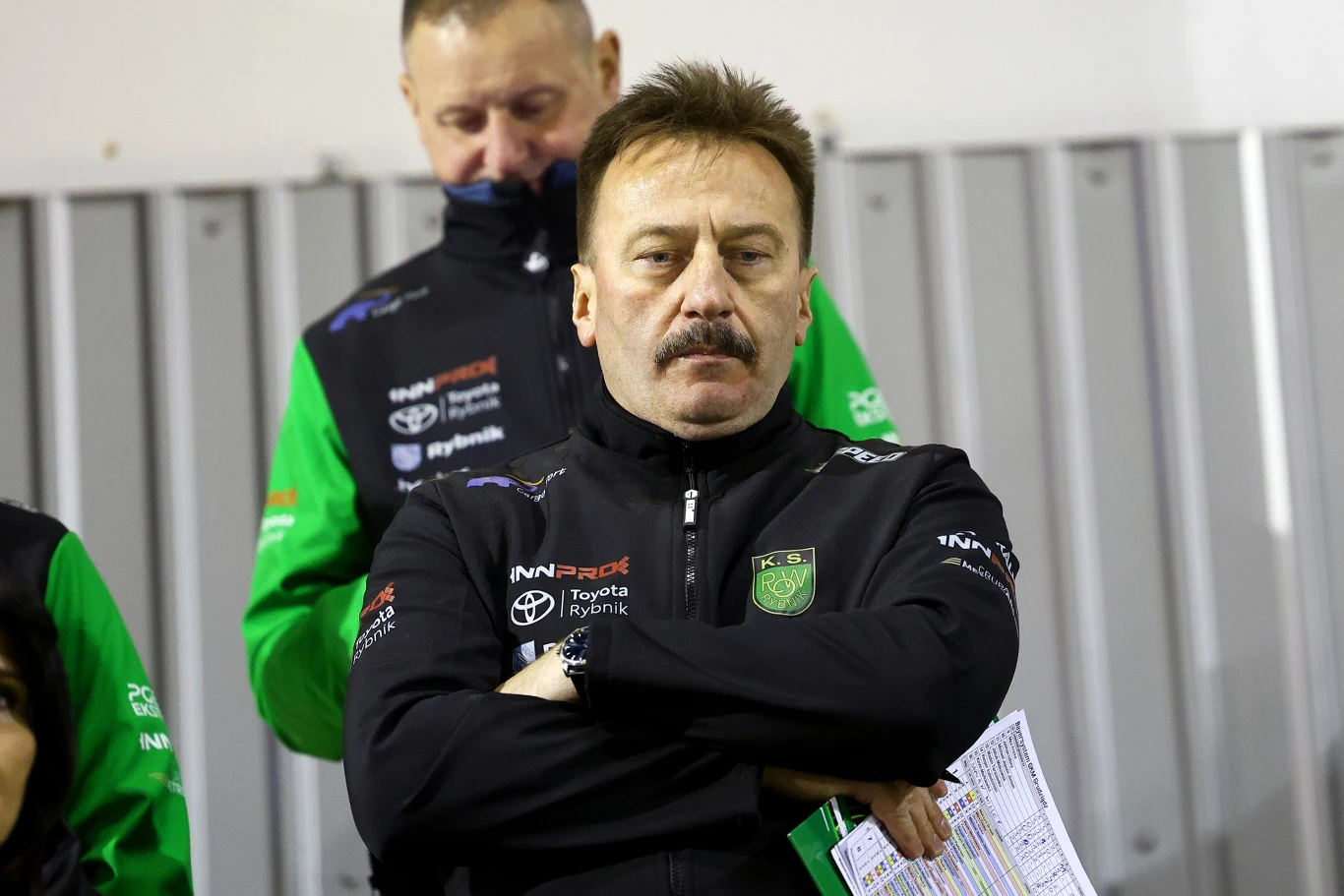 Mężczyzna z wąsem w sportowej kurtce z logotypem drużyny, trzymający złożone ręce i dokumenty, stoi na tle metalowych paneli, w tle inny mężczyzna w podobnej odzieży.