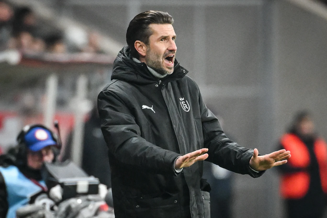 Mężczyzna w kurtce sportowej z logo klubu piłkarskiego gestykuluje na tle stadionu, najprawdopodobniej przekazując wskazówki podczas meczu.