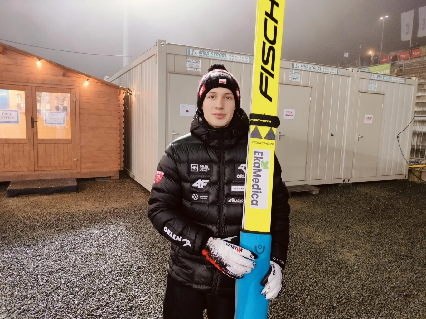 Kacper Tomasiak Młody skoczek narciarski w zimowej kurtce, rękawiczkach i czapce z logotypami sponsorów trzyma długie narty Fischer. W tle znajdują się kontenery techniczne oraz drewniany domek oświetlony lampą.
