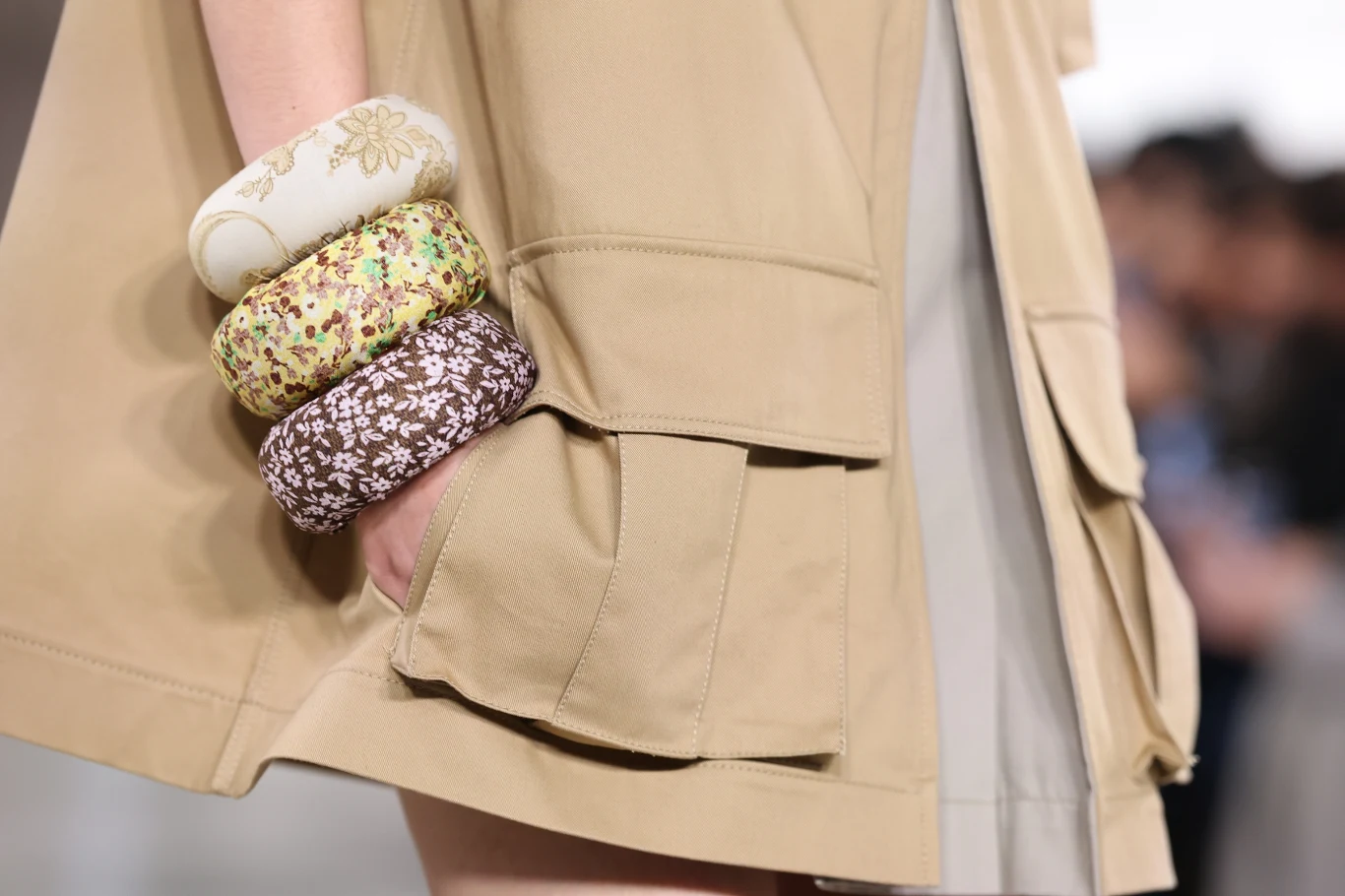 Moschino wiosna/lato 2026 Ręka osoby nosząca kilka szerokich, materiałowych bransoletek w różnych wzorach kwiatowych, opierająca się na beżowym płaszczu z dużą kieszenią.