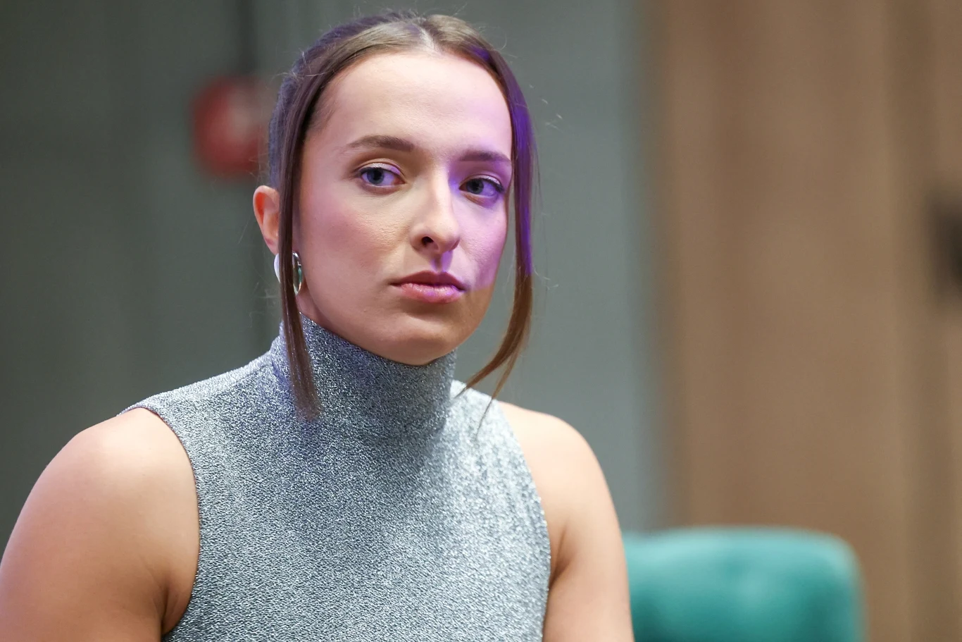 Kobieta z ciemnymi włosami spiętymi z tyłu, ubrana w szary golf bez rękawów, siedzi w pomieszczeniu z niewyraźnym tłem i patrzy w stronę aparatu z neutralnym wyrazem twarzy.