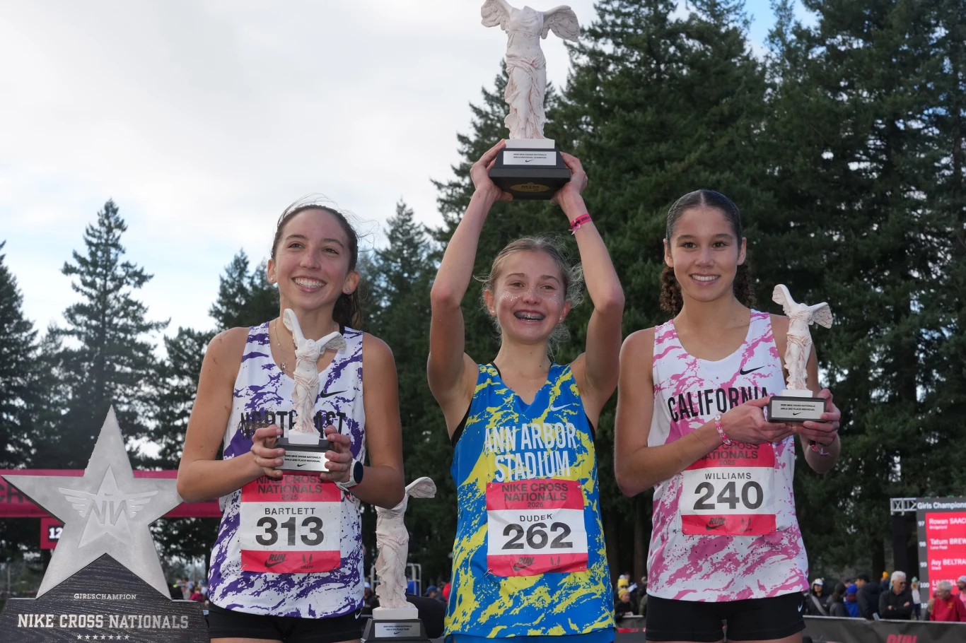 Trzy uśmiechnięte dziewczyny stoją na podium, każda trzyma statuetkę zwycięzcy, w tle widoczne drzewa oraz elementy wydarzenia sportowego.