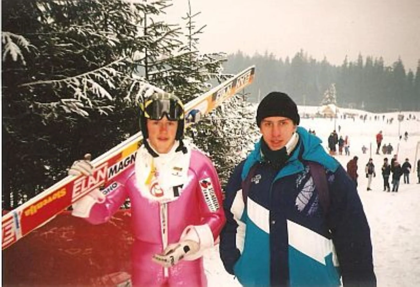 Dwóch mężczyzn pozuje na tle ośnieżonego lasu, jeden z nich ubrany jest w różowy kombinezon do skoków narciarskich i trzyma narty, drugi nosi zimową kurtkę i czapkę, w tle widać ludzi oraz stok pokryty śniegiem.