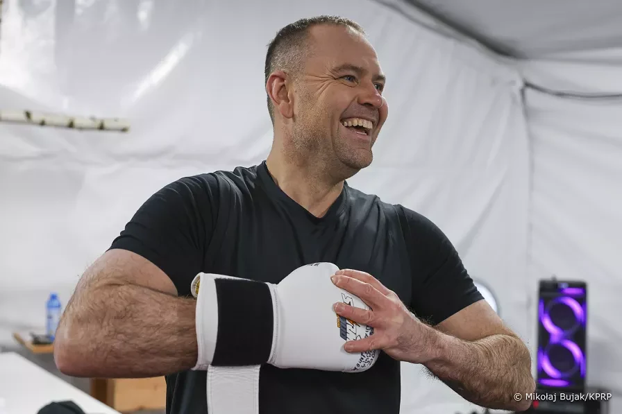 Uśmiechnięty mężczyzna w czarnej koszulce owija dłonie bandażami bokserskimi, stojąc w białym, oświetlonym pomieszczeniu.