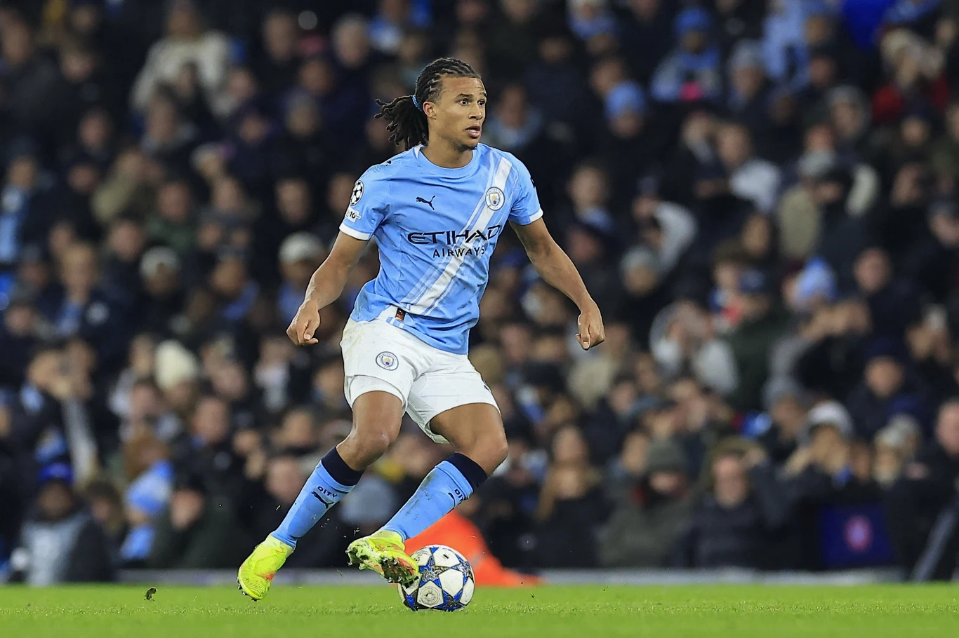 Piłkarz w niebieskim stroju Manchester City prowadzi piłkę podczas meczu, wokół niego widać rozmyte postacie kibiców na trybunach.