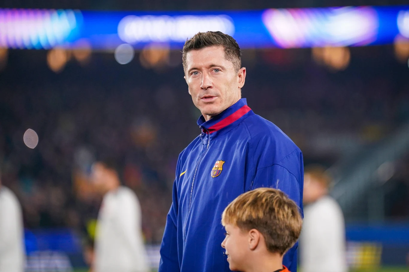 Mężczyzna w niebieskiej kurtce z herbem FC Barcelona patrzy przed siebie, w tle widoczne rozmyte postacie oraz kolorowe światła stadionu, obok niego stoi chłopiec w jasnych włosach.