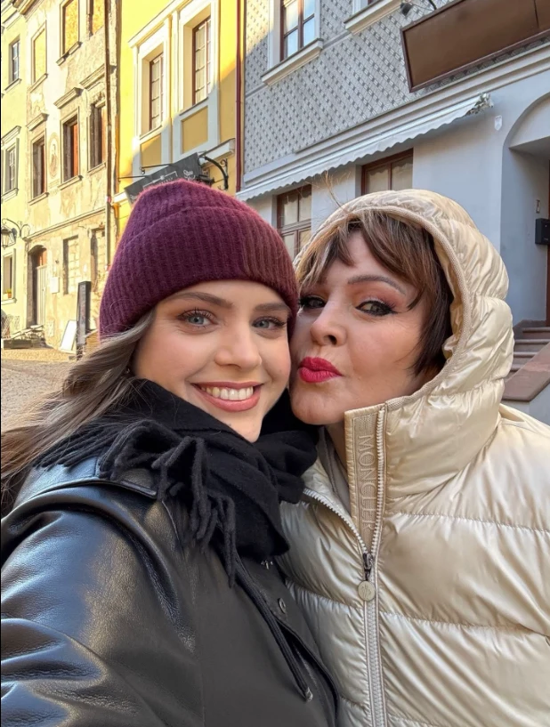 Izabela Trojanowska i jej córka Roxana Dwie kobiety ubrane w zimowe kurtki, jedna z nich nosi bordową czapkę, druga jasną kurtkę z zamkiem. Obie uśmiechnięte, stoją blisko siebie na tle kolorowych kamienic w słoneczny dzień.