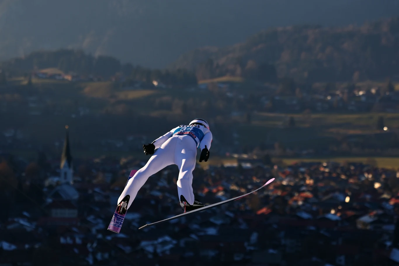 Skoczek narciarski w locie nad miastem, z rozległym widokiem na dolinę i góry w tle, w pełnym stroju sportowym i numerze startowym, dynamiczny moment w czasie zawodów.