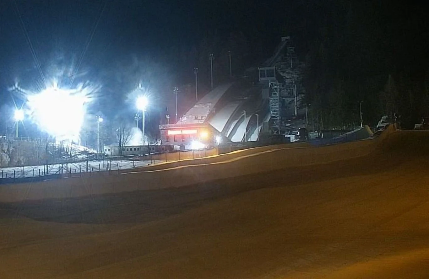 Rozświetlona skocznia narciarska nocą, wyraźnie widoczne światła oświetlające obiekt oraz trybuny i wieża sędziowska w tle, na pierwszym planie fragment igelitowej lub torowej powierzchni.