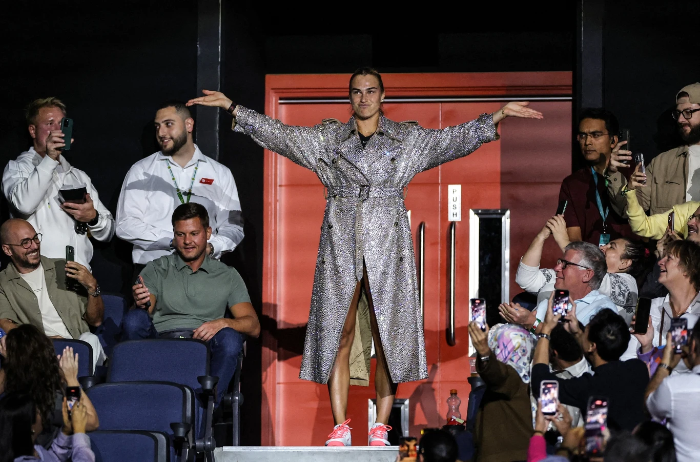Osoba w błyszczącym płaszczu i sportowych butach z uniesionymi ramionami stoi na schodach wśród publiczności, która fotografuje ją telefonami. W tle widoczna czerwona ściana i grupa wiwatujących ludzi.