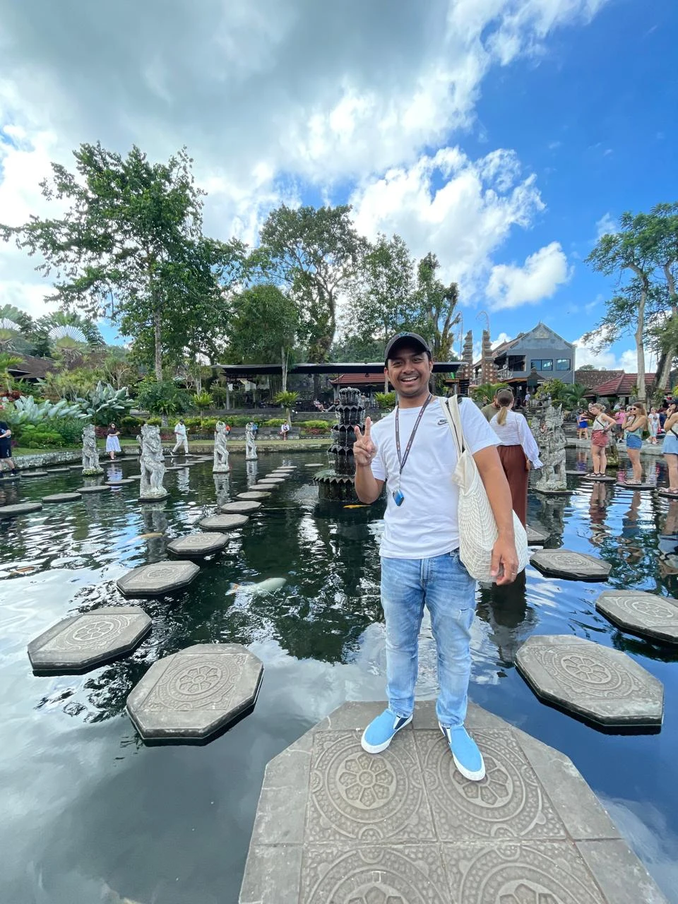 Mężczyzna w białej koszulce i czapce stoi na ozdobnej płycie kamiennej pośród stawu, wokół znajdują się inne osoby oraz ozdobna architektura ogrodowa i bujna zieleń, niebo z lekkim zachmurzeniem.