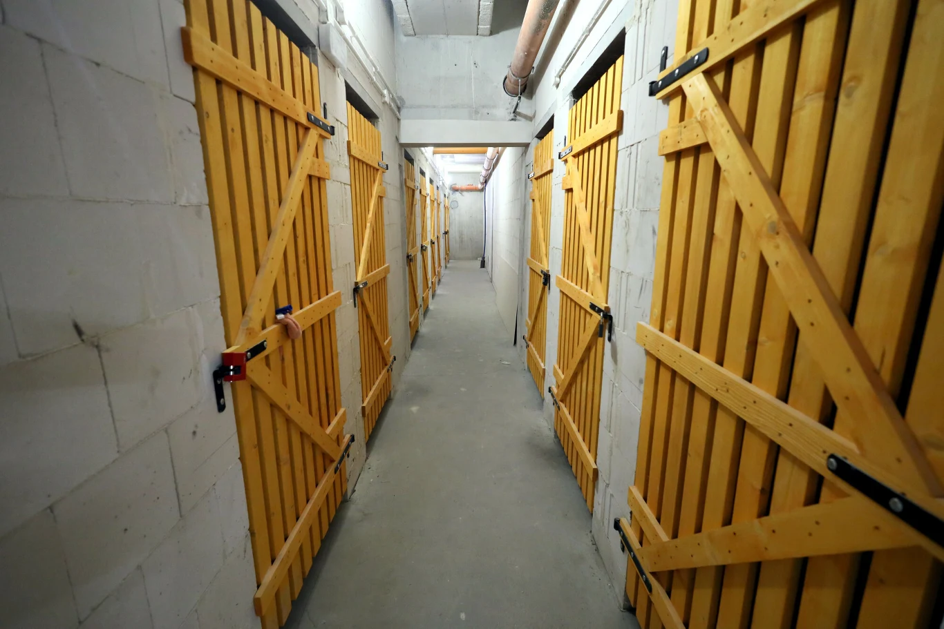 Długi, betonowy korytarz z równolegle ustawionymi drewnianymi drzwiami po obu stronach, każdy boks zamknięty kłódką, widoczne instalacje na suficie, oświetlenie sztuczne.