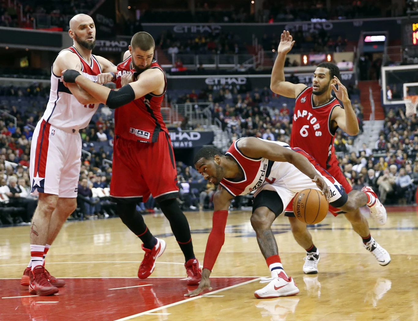 Zawodnik w białym stroju próbuje odzyskać piłkę, zbliżając się do parkietu, podczas gdy dwaj koszykarze w czerwonych strojach rywalizują z nim fizycznie, a jeden z nich blokuje przeciwnika, trzecia postać z czerwonej drużyny podnosi rękę w geście obrony