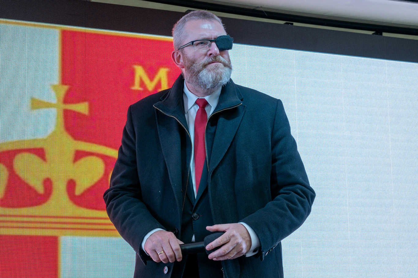 Mężczyzna z brodą i przepaską na oku stoi w eleganckim płaszczu i czerwonym krawacie na tle dużego herbu z koroną oraz czerwono-żółtą kolorystyką.