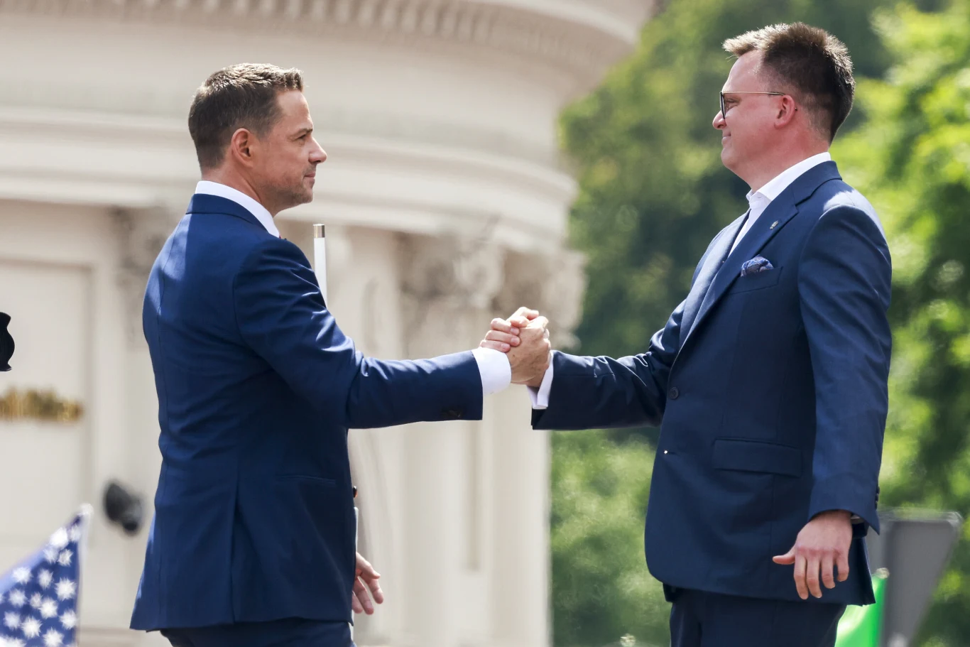 Dwóch mężczyzn w garniturach podaje sobie dłonie w geście powitania lub zawarcia porozumienia, stojąc na zewnątrz na tle jasnego budynku i zieleni.