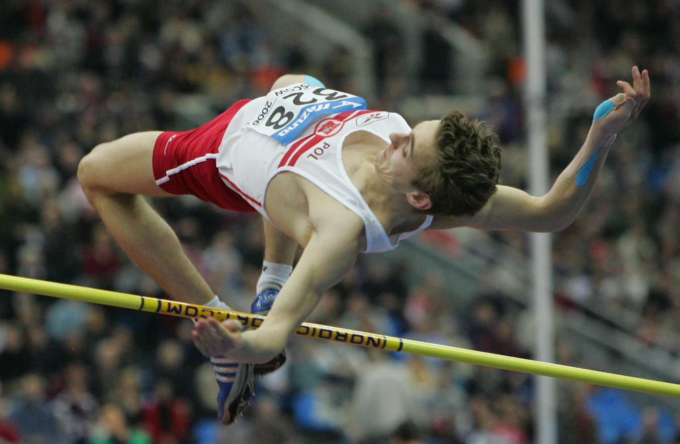 Robert Wolski Lekkoatleta w biało-czerwonym stroju przeskakuje przez poprzeczkę podczas konkurencji skoku wzwyż na stadionie sportowym, w tle publiczność na trybunach.