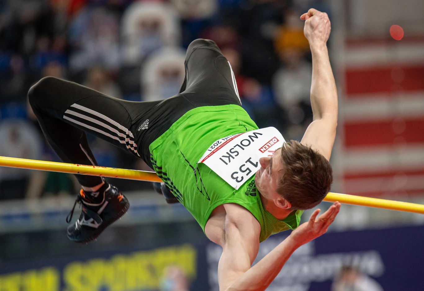 Robert Wolski Lekkoatleta w zielonej koszulce oraz numerem startowym wykonuje skok wzwyż, unosząc się nad poprzeczką na tle sportowej hali.