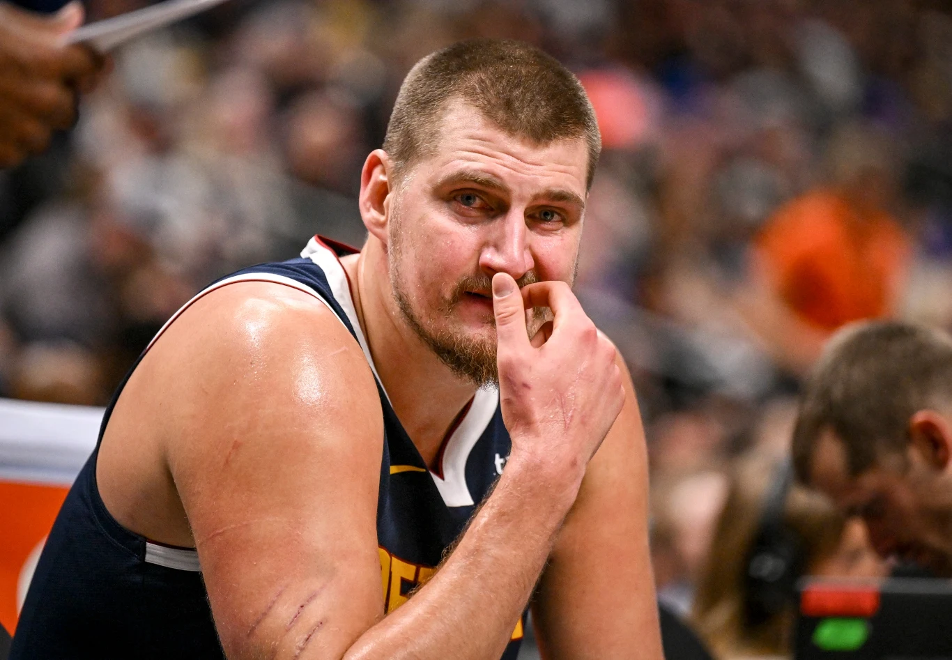 Koszykarz w sportowym stroju siedzi na ławce podczas meczu, opiera rękę o twarz, jego wyraz twarzy sugeruje zmęczenie lub skupienie, w tle widoczna rozmyta publiczność.