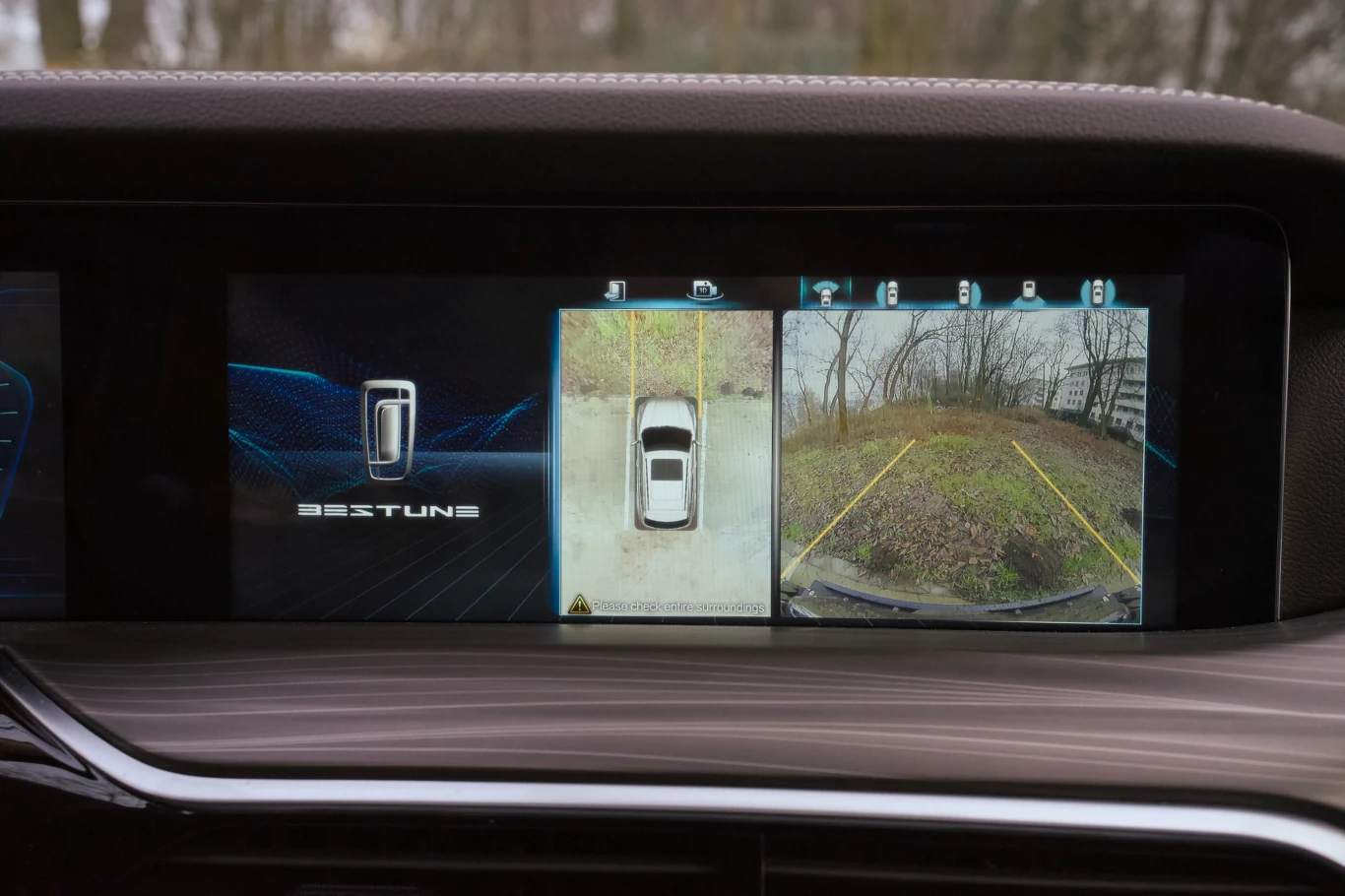 Centralny wyświetlacz samochodu pokazuje obraz z kamer cofania oraz widok z góry na pojazd, wraz z liniami pomocniczymi ułatwiającymi parkowanie. Otoczenie pojazdu to trawiasty i asfaltowy teren na tle drzew i budynków.