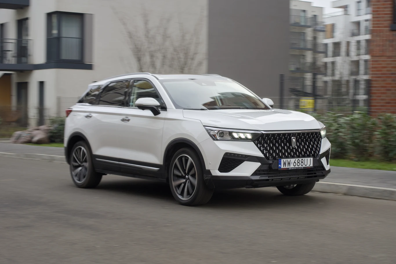 Biały nowoczesny SUV poruszający się po miejskiej ulicy, na tle współczesnej zabudowy mieszkaniowej.