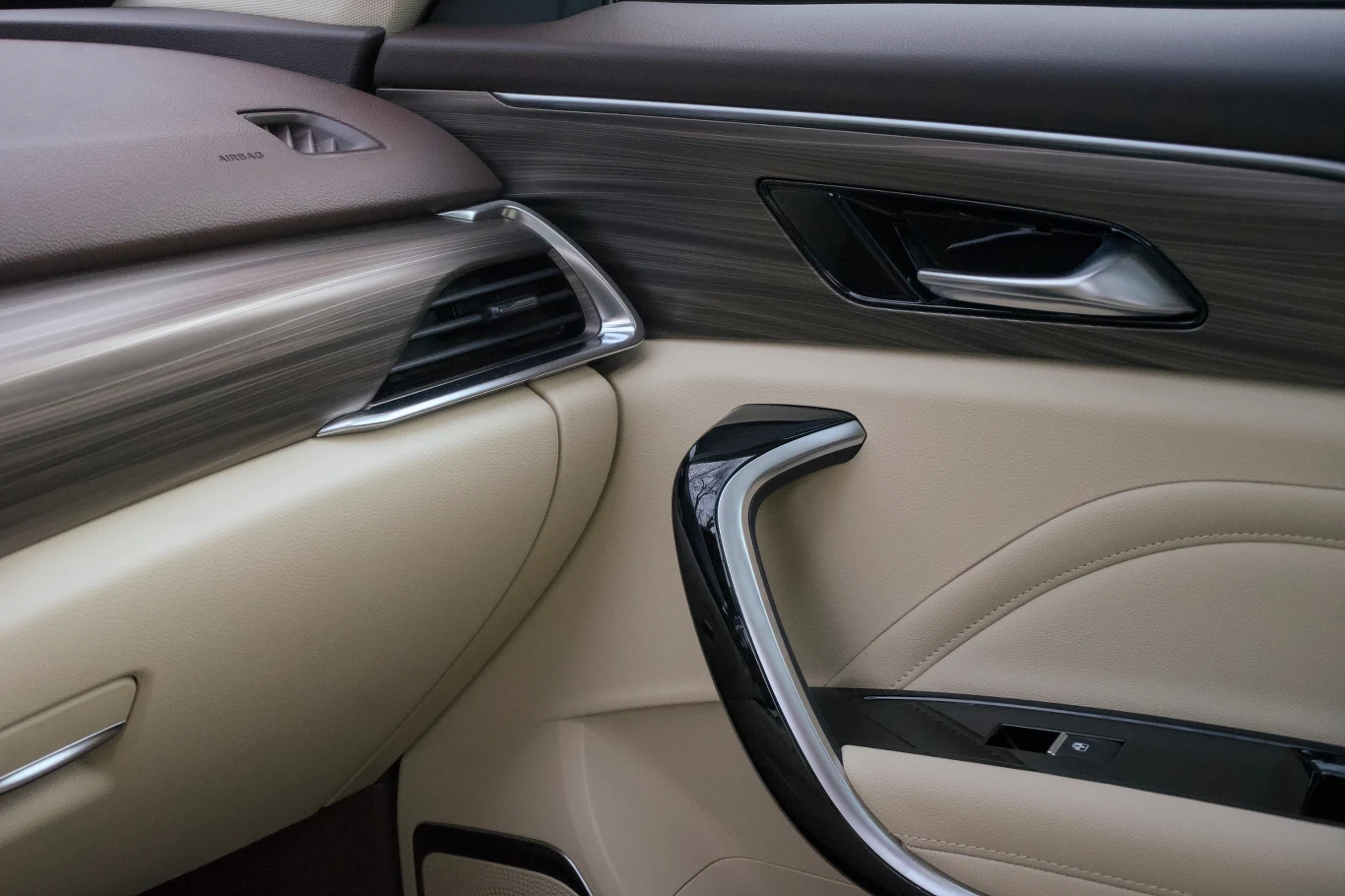 Luksusowe wnętrze samochodu, fragment drzwi wykończony jasną skórą i eleganckimi, metalowymi oraz drewnopodobnymi elementami dekoracyjnymi, widoczne są także nawiew i klamka.