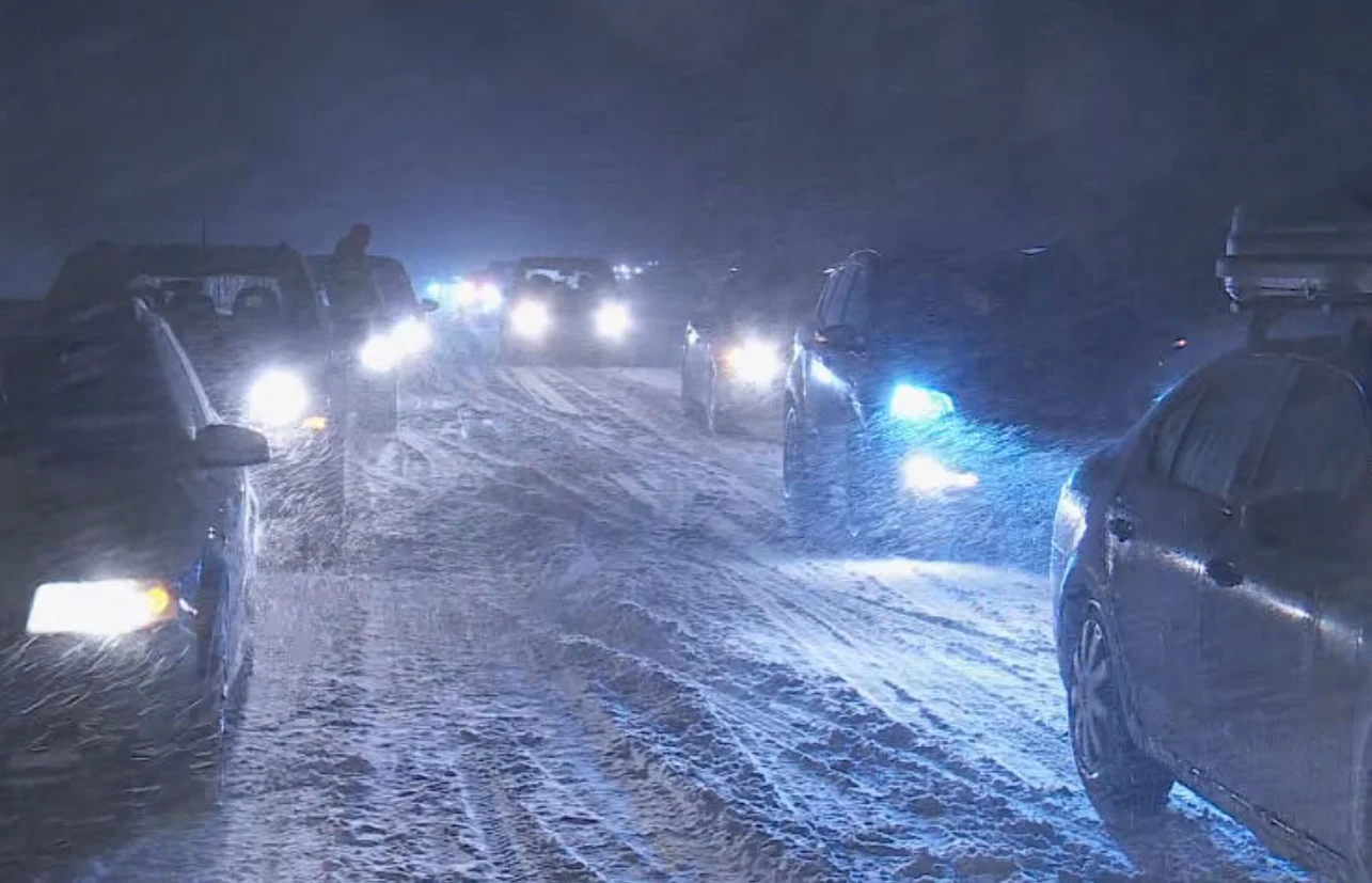Szereg samochodów jadących w nocy podczas intensywnej zamieci śnieżnej. Wiązki reflektorów oświetlają wirujący śnieg, droga zasypana białym puchem utrudnia widoczność i poruszanie się pojazdów.