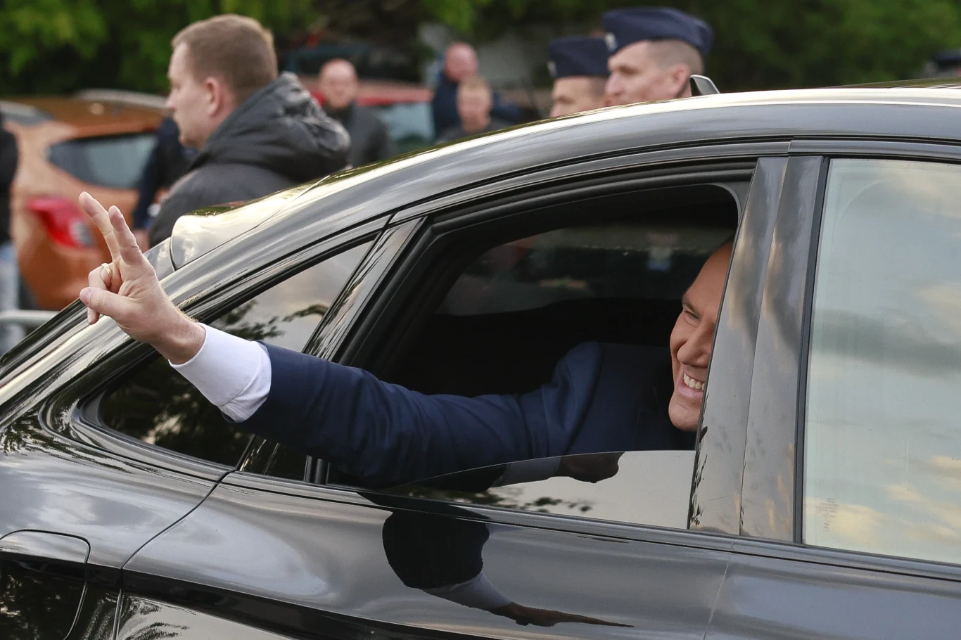 Mężczyzna w garniturze pokazuje znak zwycięstwa przez opuszczoną szybę czarnego samochodu, uśmiechając się. W tle widoczne są inne osoby oraz kilka pojazdów.