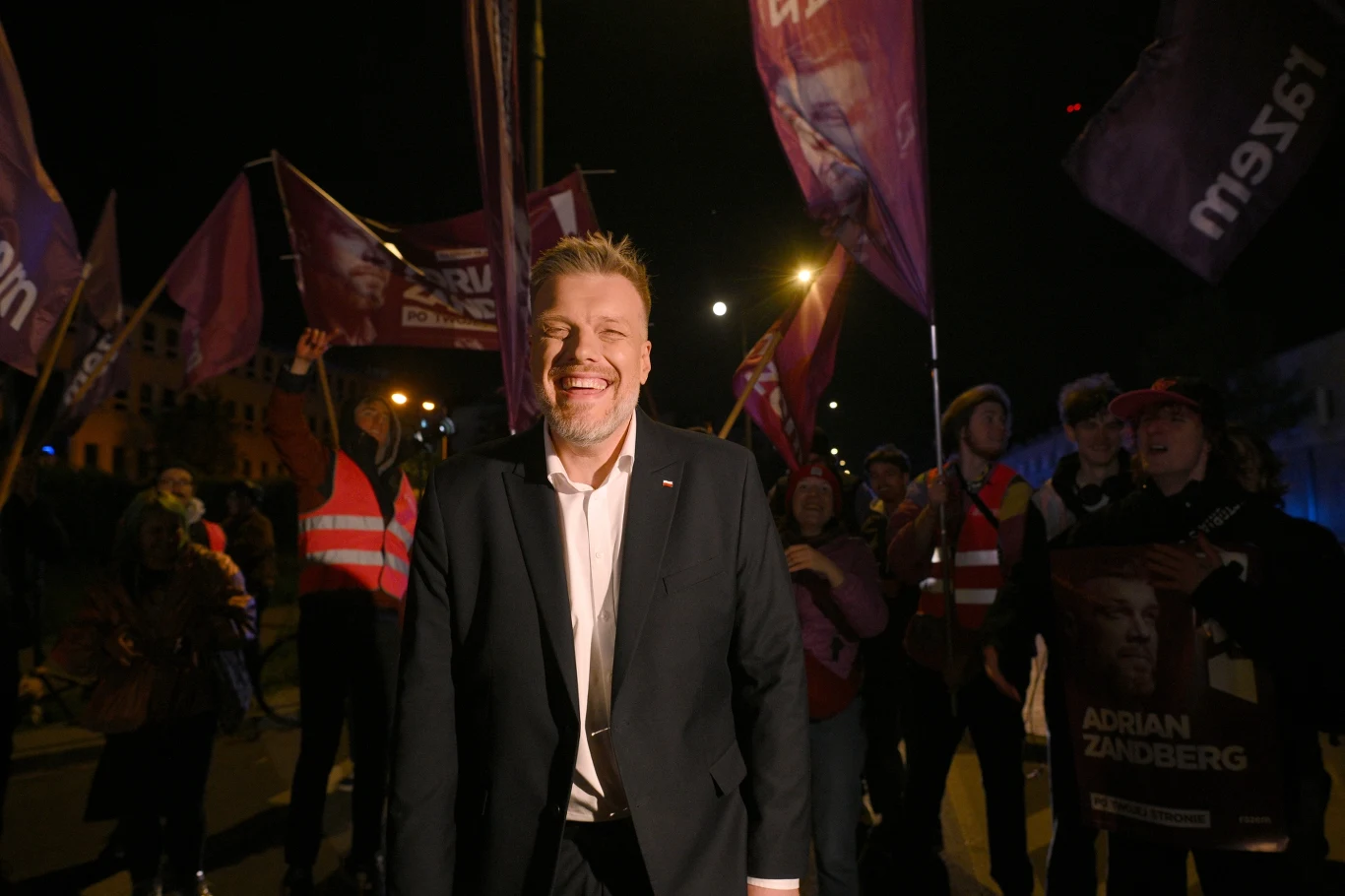 Mężczyzna w garniturze uśmiecha się, stojąc na pierwszym planie wśród grupy ludzi z flagami partyjnymi i plakatami w tle podczas wieczornego zgromadzenia ulicznego.
