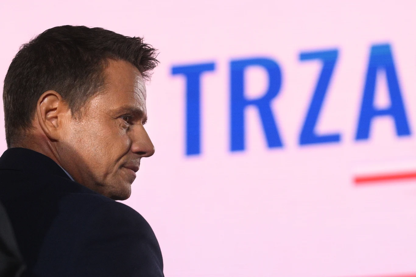 Mężczyzna w średnim wieku o poważnym wyrazie twarzy na tle jasnego ekranu z wyraźnym napisem, który kończy się na litery 'TRZA'.