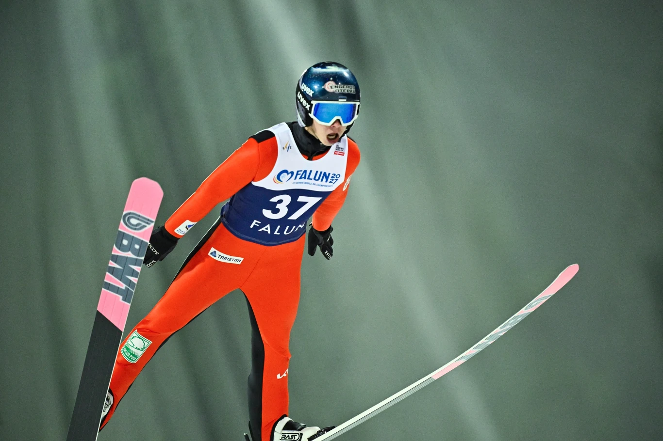 Artti Aigro Zawodnik w czerwonym kombinezonie wykonuje skok narciarski w trakcie zawodów, lecąc w powietrzu na rozstawionych nartach, w tle widoczny rozmyty śnieżny krajobraz.